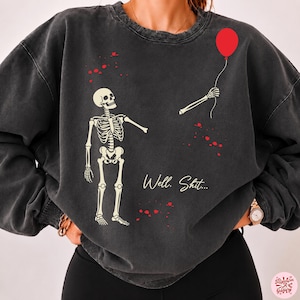 Könnte beinhalten: Ein anthrazitfarbenes Sweatshirt mit Rundhalsausschnitt und einer Grafik eines Skeletts, das auf seinen eigenen abgetrennten Arm mit einem roten Luftballon blickt. Der Text "Well. Shit..." steht in weißer Schreibschrift darunter.