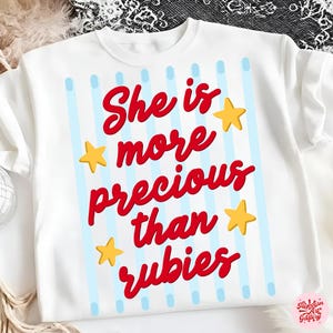 Puede incluir: Sudadera blanca con el texto rojo "She is more precious than rubies". El diseño incluye rayas azules claras y estrellas amarillas. Una parte superior cómoda e informal.