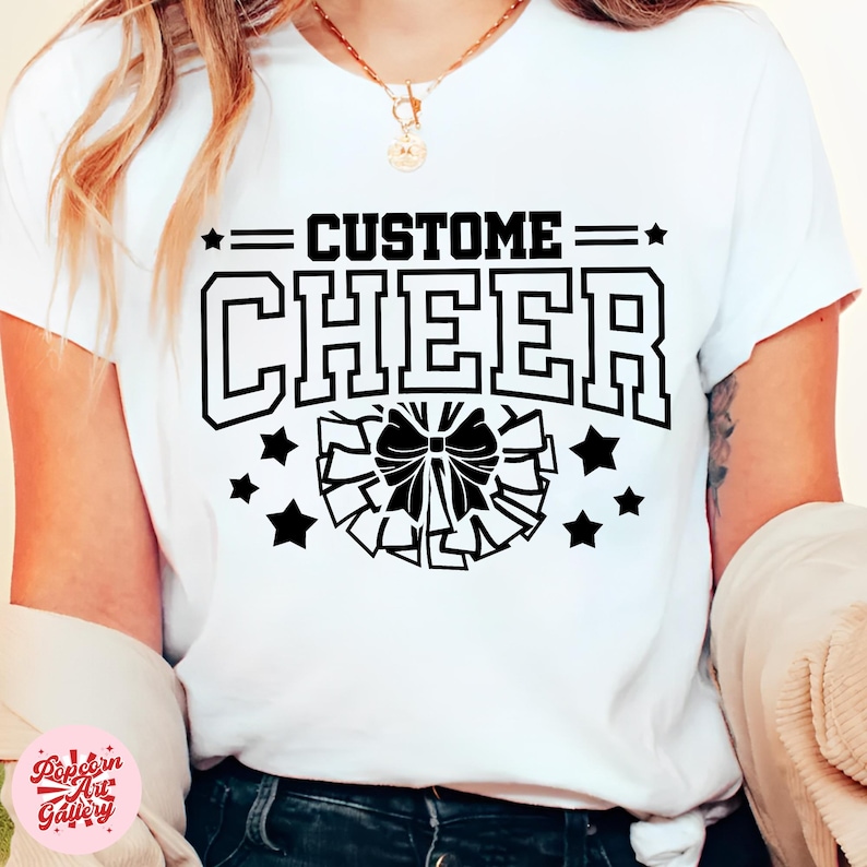 May include: White t-shirt with the text "CUSTOM CHEER" in a black, collegiate-style font, with a black bow and star graphic. The shirt is a crew neck and appears to be made of a soft material.