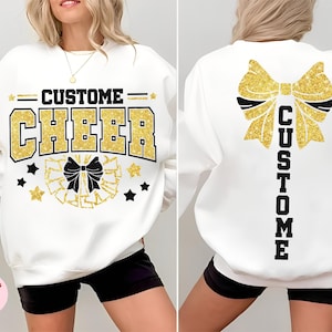 May include: White sweatshirt with "CUSTOM CHEER" in gold glitter lettering. The front features a bow and stars, while the back displays a large bow and the word "CUSTOM". A cheerleading-themed design.
