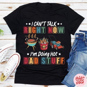Può includere: T-shirt nero con la scritta "I CAN'T TALK RIGHT NOW I'M DOING HOT DAD STUFF" a blocchi colorati. La grafica include una griglia con cibo, un secchio di bevande e un tosaerba. Una maglietta divertente e casual.