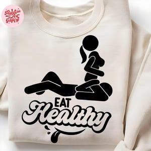 Peut inclure: Sweat-shirt crème avec un graphisme noir de deux personnages stylisés dans une position sexuelle, avec les mots "EAT Healthy" dans une police stylisée. Le sweat-shirt a un col rond et des manches longues.