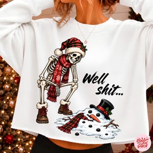 May include: A white long-sleeve sweatshirt featuring a graphic of a skeleton wearing a red plaid Santa hat, scarf, and brown boots, standing over a melted snowman. The text "Well shit..." is printed in black script next to the skeleton.