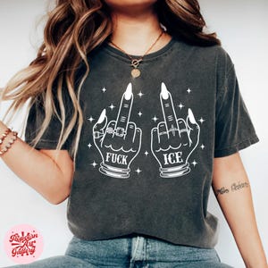 Peut inclure: T-shirt gris fonc&eacute; avec un motif blanc de deux mains faisant un doigt d'honneur. Le mot "FUCK" est en dessous d'une main et "ICE" en dessous de l'autre. Les mains sont orn&eacute;es de bagues et de longs ongles blancs.