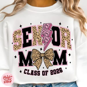 Puede incluir: Sudadera blanca con las palabras "SENIOR MOM" en negro y estampado de leopardo, con un rayo rosa y un lazo. El texto "CLASS OF 2026" está debajo del lazo, con detalles de estrellas negras.
