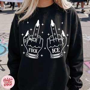Peut inclure: Sweat-shirt noir avec un motif blanc de deux mains faisant un doigt d'honneur. Le mot "FUCK" est imprim&eacute; sous une main, et "ICE" sous l'autre. Les mains sont orn&eacute;es de bagues et de longs ongles, avec des &eacute;toiles.