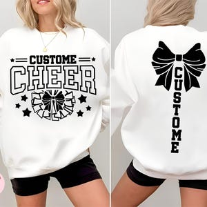 May include: White sweatshirt with the words "CUSTOM CHEER" in black block letters. The front features a cheer bow and stars. The back of the sweatshirt has a large bow and the word "CUSTOM" vertically.