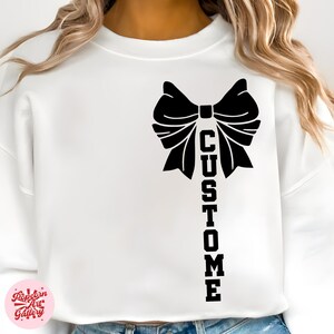 May include: White sweatshirt featuring a large black bow design with the word "CUSTOM" printed vertically below it. The sweatshirt is a solid white color, and the design is centered on the front.