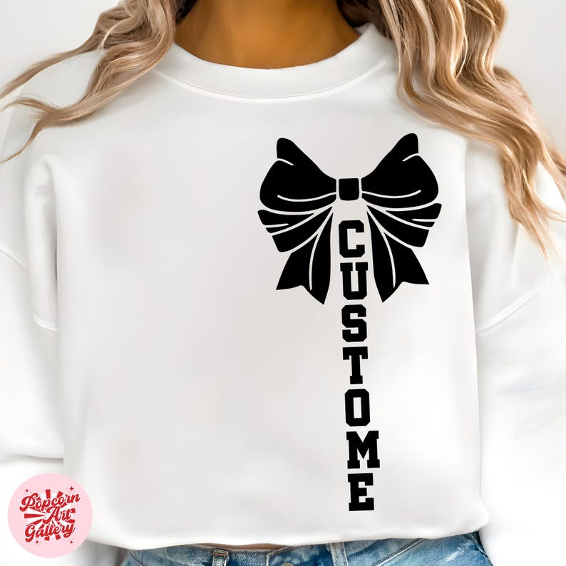 May include: White sweatshirt featuring a large black bow design with the word "CUSTOM" printed vertically below it. The sweatshirt is a solid white color, and the design is centered on the front.