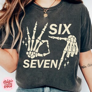 May include: Dark gray t-shirt with a cream-colored skeleton hand graphic forming the numbers six and seven. The word "SIX" is above the hand gesture, and "SEVEN" is below. Sparkle accents are included.