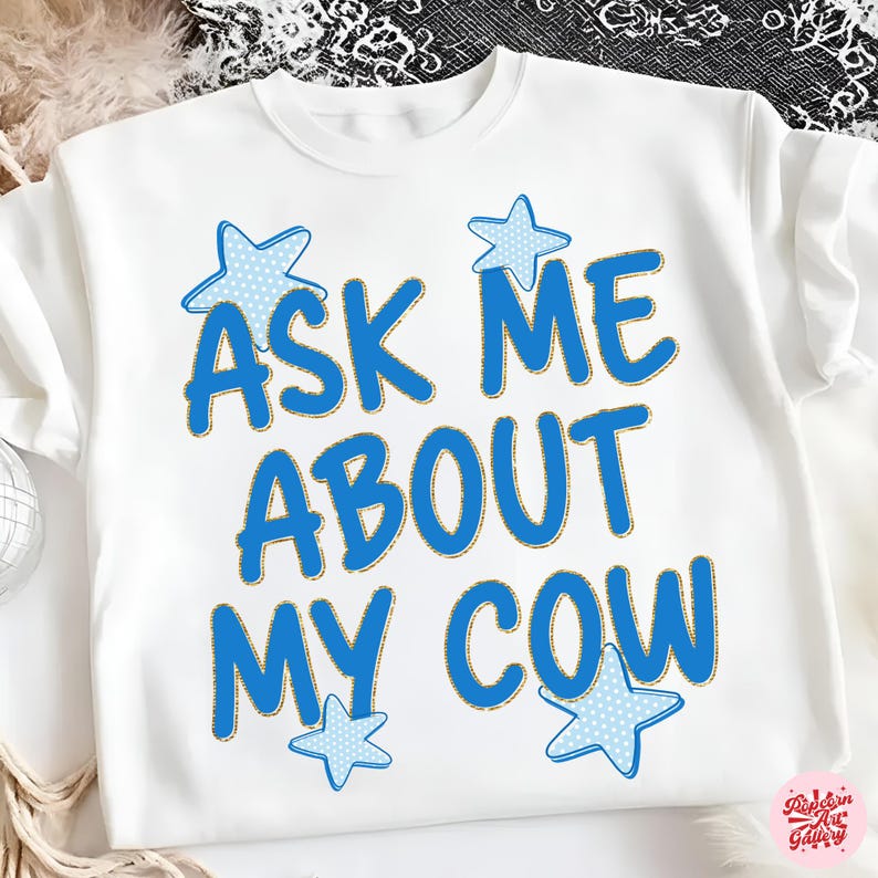 May include: White sweatshirt with the text "ASK ME ABOUT MY COW" in blue, outlined in gold. Blue star accents with white polka dots are placed around the text. The sweatshirt is a casual, comfortable style.