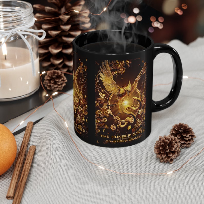 A black mug with an image of the Ballad of Songbirds and Snakes symbol printed on it. Steam is rising from the mug which as a dark liquid inside of it. The mug sits on a white cloth with small pinecones next to it. A candle and larger pinecone sit in the background. Cinnamon sticks sit off to the side of the mug.