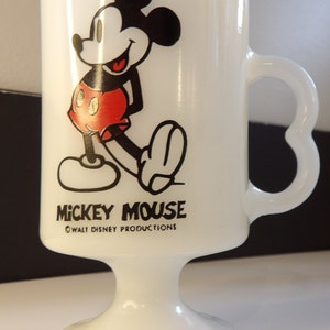 May include: White ceramic mug with a black and red image of Mickey Mouse. The mug has a heart-shaped handle and a pedestal base. The text "Mickey Mouse" and "&copy; WALT DISNEY PRODUCTIONS" is printed on the mug.