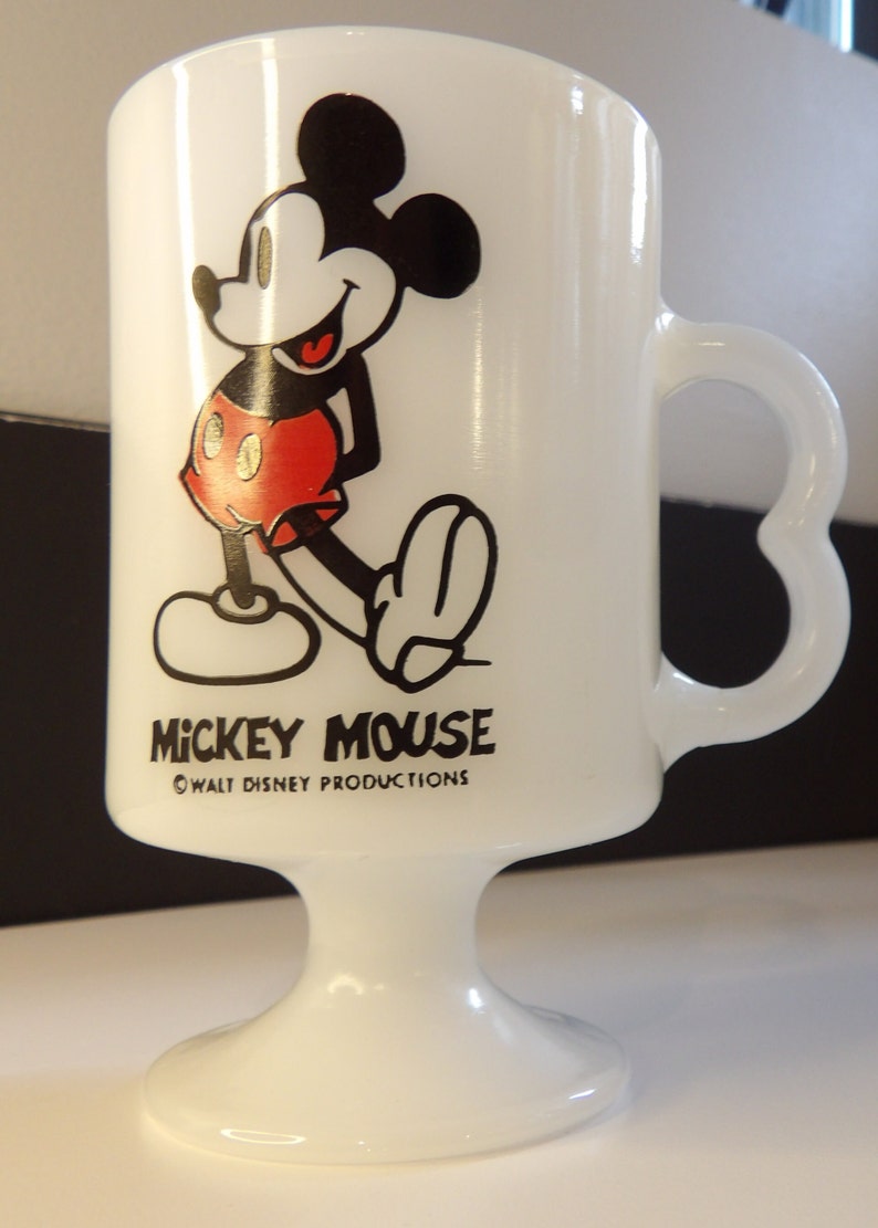 May include: White ceramic mug with a black and red image of Mickey Mouse. The mug has a heart-shaped handle and a pedestal base. The text "Mickey Mouse" and "&copy; WALT DISNEY PRODUCTIONS" is printed on the mug.