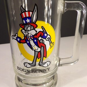 May include: A glass Bugs Bunny mug with a red, white, and blue illustration of Bugs Bunny dressed as Uncle Sam. The mug has a yellow circle around Bugs Bunny and the text "Bugs Bunny" at the bottom. The text "Warner Bros. Inc. 1975" is also at the bottom of the mug.
