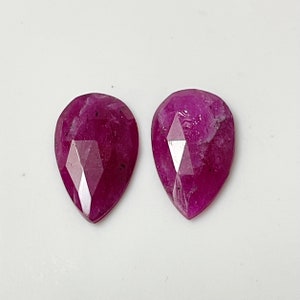 May include: Two pear-shaped faceted ruby gemstones, each with a deep pink hue and a slightly cloudy appearance.