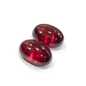 Pu&ograve; includere: Due pietre preziose rosso scuro di forma ovale con una finitura cabochon liscia e lucida. Le pietre mostrano una qualit&agrave; traslucida con riflessi di luce interni visibili e una consistenza superficiale lucida su uno sfondo bianco.