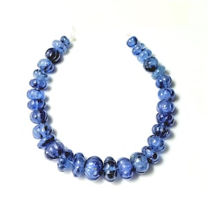 May include: A strand of blue gemstone beads, each bead is faceted and has a slightly irregular shape. The beads are strung on a white thread and are arranged in a circle.