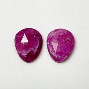 May include: Two pink faceted gemstone cabochons with a triangular facet in the center. The stones are a light pink color with white inclusions.