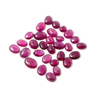 May include: A collection of 25 oval-shaped, faceted, pink gemstones. The gemstones are scattered on a white background.