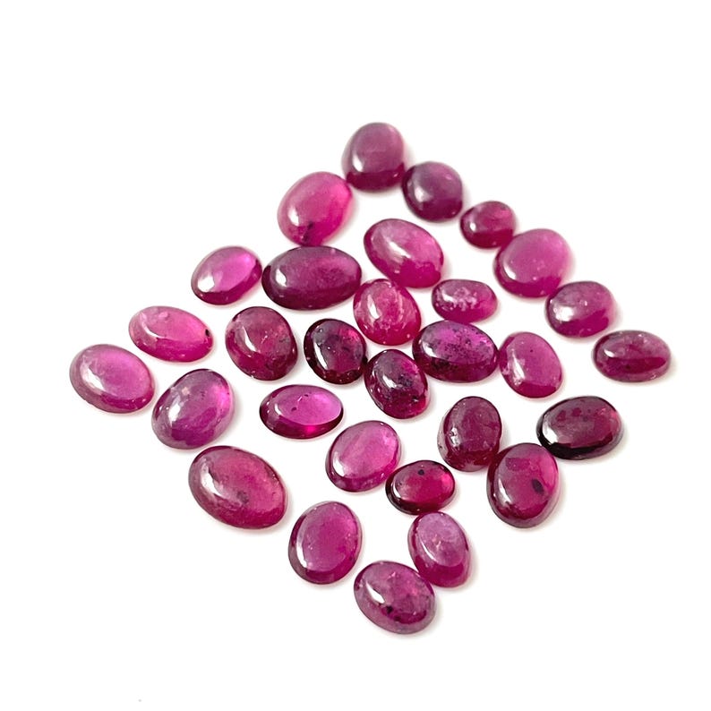 May include: A collection of 25 oval-shaped, faceted, pink gemstones. The gemstones are scattered on a white background.
