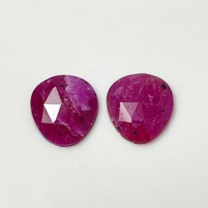 May include: Two pink ruby gemstones with a faceted cut. The gemstones are heart-shaped and have a dark pink color.