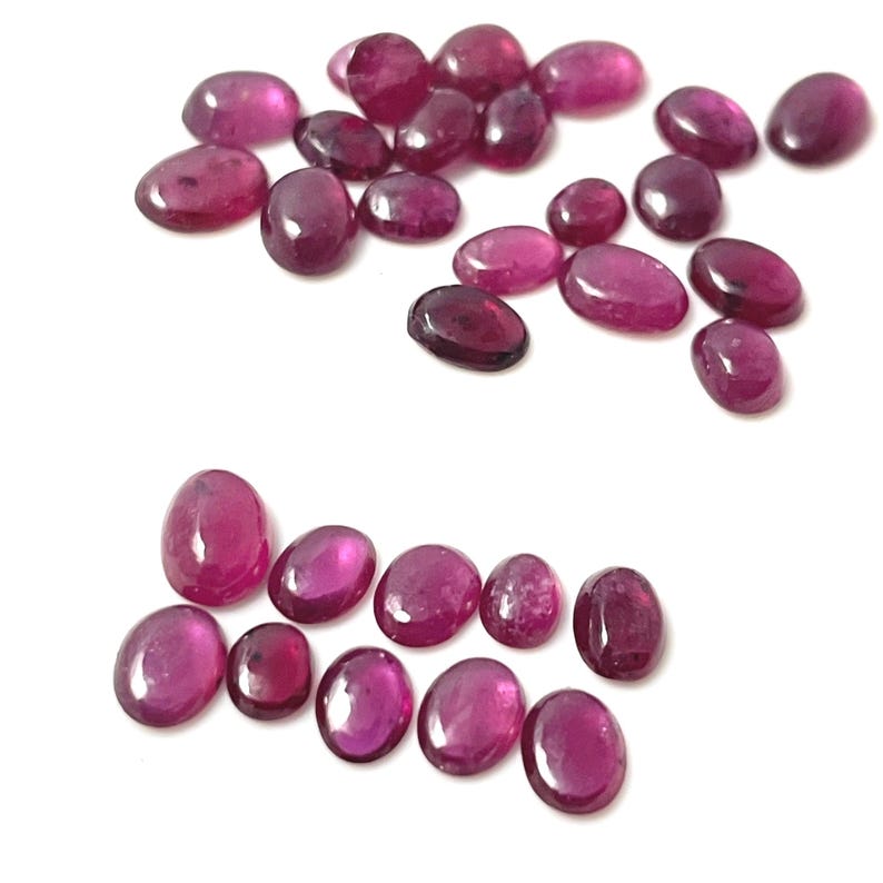 May include: A collection of oval-shaped, deep pink gemstones. The gemstones are scattered on a white surface.