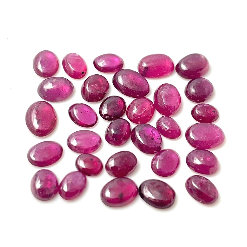 May include: A collection of 25 oval-shaped, faceted, ruby red gemstones. The stones are various sizes and have a polished finish.