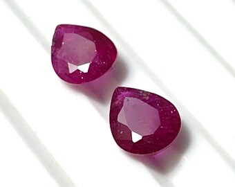 May include: Two pear-shaped ruby gemstones, each with a deep red hue and a faceted cut. The gemstones are set against a white background.