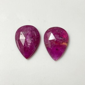 May include: Two pear-shaped pink gemstones with a faceted cut. The gemstones have a dark pink hue with a slight purple tint.