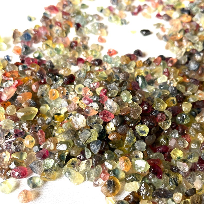 May include: A large pile of colorful, faceted gemstones. The gemstones are various shades of red, blue, green, yellow, and brown. The gemstones are small and appear to be rough cut.