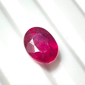 Puede incluir: Una gema ovalada de color rojo vibrante con un corte facetado. La gema tiene un ligero tono rosado y está colocada sobre un fondo blanco.