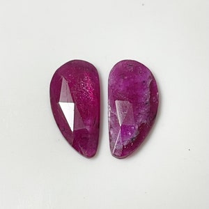 May include: Two faceted, teardrop-shaped gemstones with a deep pink hue. The stones have a slightly irregular shape and a polished finish.