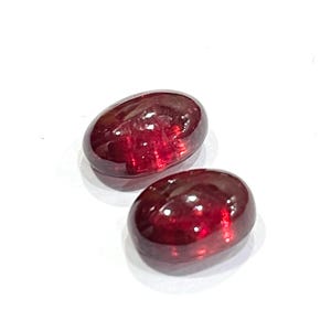 Pu&ograve; includere: Due pietre preziose rosso scuro di forma ovale con un taglio cabochon liscio e levigato. Le pietre mostrano una qualit&agrave; traslucida con riflessi di luce interni e una finitura superficiale lucida, che mette in risalto un colore cremisi ricco e saturo.
