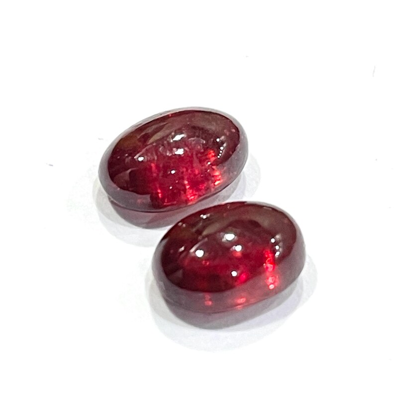 Pu&ograve; includere: Due pietre preziose rosso scuro di forma ovale con un taglio cabochon liscio e levigato. Le pietre mostrano una qualit&agrave; traslucida con riflessi di luce interni e una finitura superficiale lucida, che mette in risalto un colore cremisi ricco e saturo.