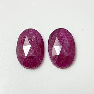 May include: Two oval-shaped ruby gemstones with a faceted cut. The rubies are a deep pink color with a slight sparkle.
