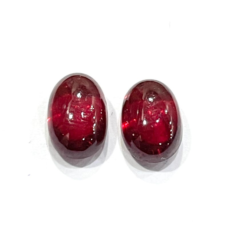 Pu&ograve; includere: Due pietre preziose di colore rosso intenso di forma ovale con una finitura cabochon liscia e lucida. Le pietre mostrano una qualit&agrave; traslucida con inclusioni interne e riflessi di luce sulle superfici arrotondate, mettendo in mostra una ricca tonalit&agrave; cremisi.