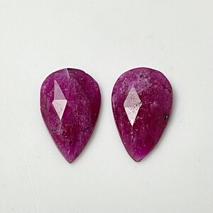 May include: Two pear-shaped pink gemstones with a faceted design. The gemstones are set in a clear, faceted setting.