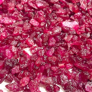 以下が含まれることがあります： 不規則な形をした小さなルビーレッドの宝石が大量に散らばっているクローズアップ。宝石は白い表面に散らばっています。