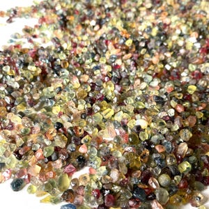 May include: A large collection of small, rough, multi-colored gemstones. The gemstones are a mix of blues, greens, yellows, and reds.