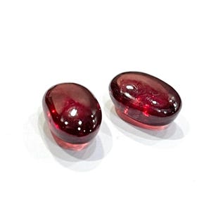 Pu&ograve; includere: Due pietre preziose traslucide di colore rosso intenso, di forma ovale e con una finitura cabochon liscia e lucida. Queste pietre presentano una ricca tonalit&agrave; cremisi scuro e una superficie lucida che riflette la luce, esaltando la forma.