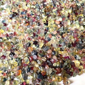 May include: A large collection of loose, faceted gemstones in various colors, including red, blue, green, yellow, and purple. The gemstones are small and irregularly shaped, and they are scattered across a white surface.