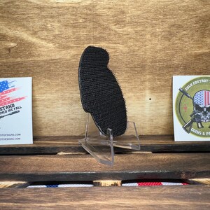 May include: Black hook and loop patch with a rough texture. The patch is on a clear plastic stand. The stand is on a wooden surface. There are two stickers in the background. One sticker has a red, white, and blue American flag with the text "United We Stand Disarmed We Fall" and a website address. The other sticker has a green background with a skull wearing a red, white, and blue American flag bandana. The skull is holding two crossed rifles. The text "Zulu Foxtrot Tactical Coins & Patches" is below the skull.