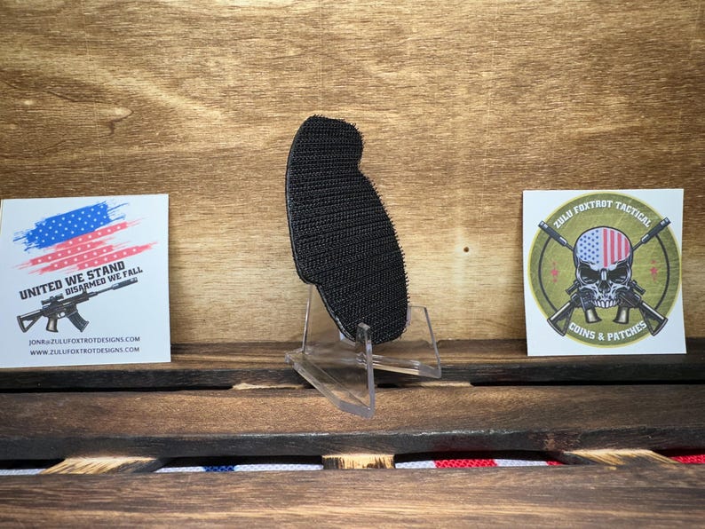 May include: Black hook and loop patch with a rough texture. The patch is on a clear plastic stand. The stand is on a wooden surface. There are two stickers in the background. One sticker has a red, white, and blue American flag with the text "United We Stand Disarmed We Fall" and a website address. The other sticker has a green background with a skull wearing a red, white, and blue American flag bandana. The skull is holding two crossed rifles. The text "Zulu Foxtrot Tactical Coins & Patches" is below the skull.