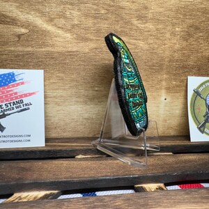 May include: A green and black embroidered patch with the text "Improvise Adapt Overcome" in gold lettering. The patch is displayed on a clear acrylic stand.