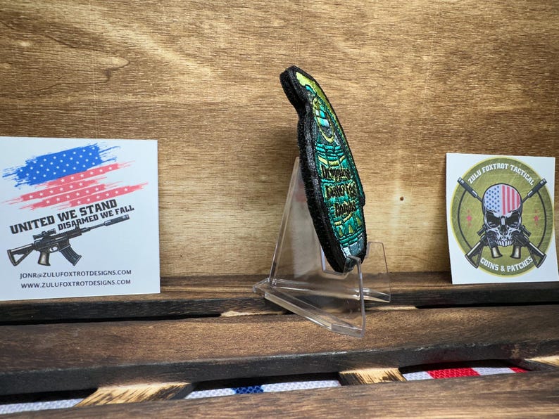 May include: A green and black embroidered patch with the text "Improvise Adapt Overcome" in gold lettering. The patch is displayed on a clear acrylic stand.