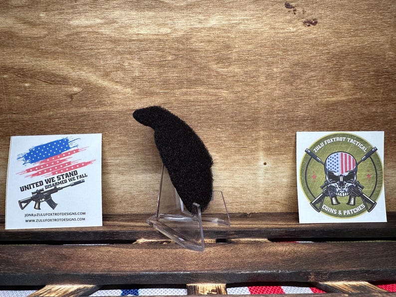 May include: A black patch with a fuzzy texture, displayed on a clear plastic stand. The patch features the text "ZULU FOXTROT TACTICAL" and a skull with an American flag design. The text "COINS & PATCHES" is also visible on the patch.