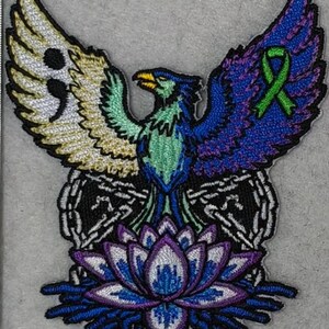 May include: Embroidered patch featuring a phoenix with outstretched wings. The wings are divided into sections with a semicolon, a green ribbon, and a mix of colors. A lotus flower is at the bottom. The patch is a mix of blue, green, purple, gold, and white.