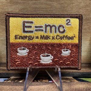 May include: A brown and yellow embroidered patch with the text "E=mc²" and "Energy = Milk x Coffee²". The patch features three small embroidered coffee cups on a brown background.