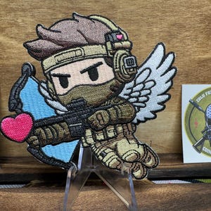 May include: Embroidered patch of a cartoon soldier with angel wings, holding a bow and arrow with a pink heart. The soldier is wearing a brown and green camouflage uniform and a black helmet with a pink heart on it. The patch is on a wooden background.